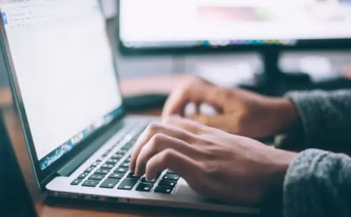 A person typing on a laptop keyboard at a wooden desk. Two computer monitors are in the background, displaying blurry screens. The person is wearing a grey sweater, and the room is softly lit.