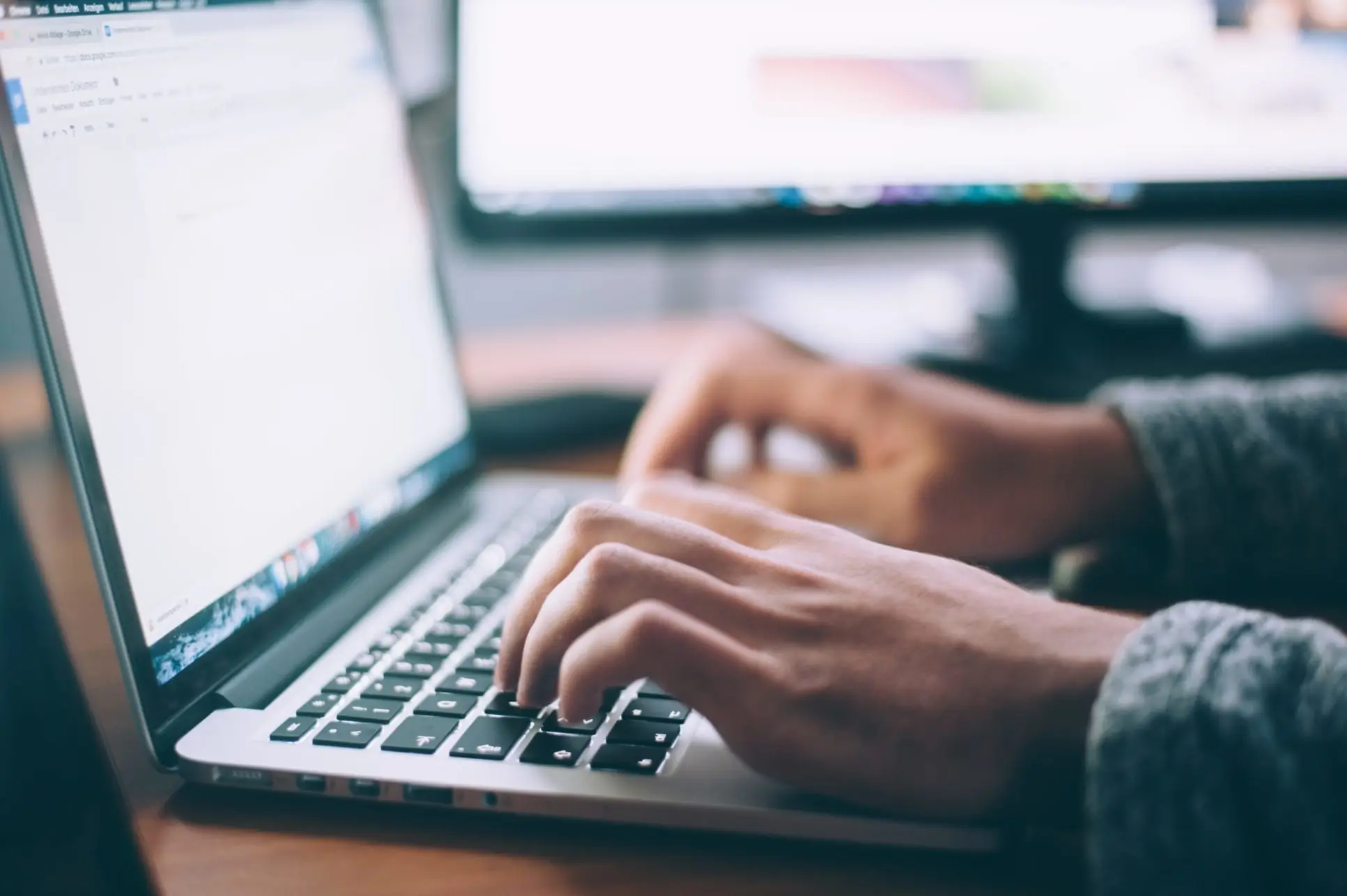 A person typing on a laptop keyboard at a wooden desk. Two computer monitors are in the background, displaying blurry screens. The person is wearing a grey sweater, and the room is softly lit.