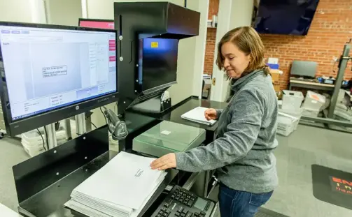 A woman in a gray sweater stands at a machine, placing white envelopes into a drop slot. A computer monitor displays a tracking interface. The room has brick walls and various office equipment and supplies.