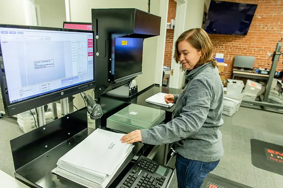 A woman in a gray sweater stands at a machine, placing white envelopes into a drop slot. A computer monitor displays a tracking interface. The room has brick walls and various office equipment and supplies.