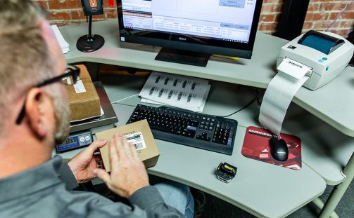 Looking down on a desk with keyboard, mouse, a label printer, scale, ring scanner, monitor, and a man applying a label to a parcel