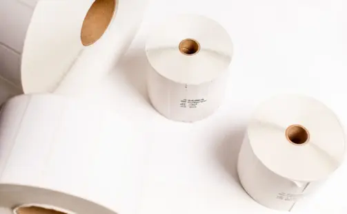 Four large rolls of white label paper on a white surface. The rolls are unrolled slightly, revealing the brown cardboard cores. Two rolls are upright, while the other two are lying horizontally, showcasing their width and thickness.