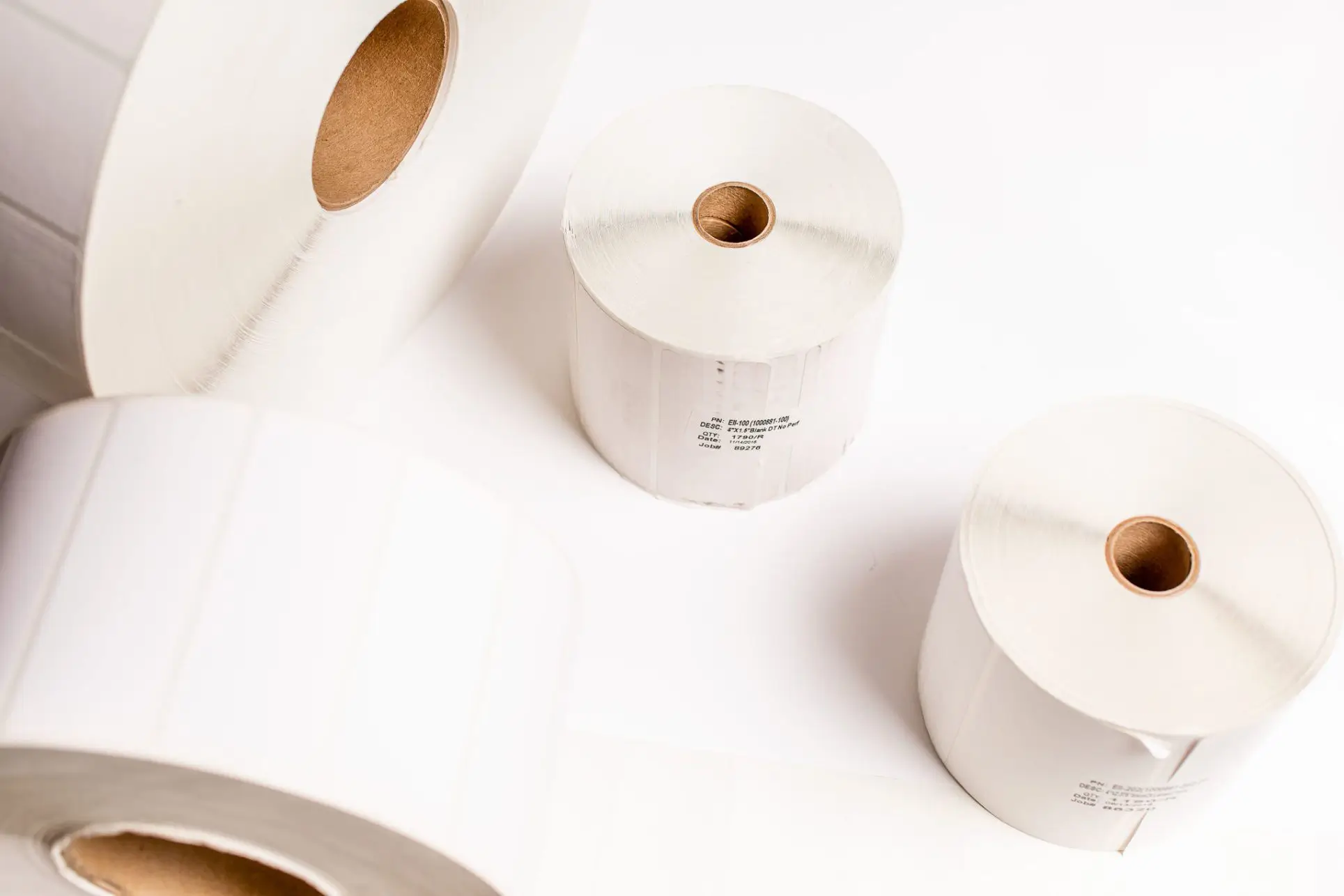 Four large rolls of white label paper on a white surface. The rolls are unrolled slightly, revealing the brown cardboard cores. Two rolls are upright, while the other two are lying horizontally, showcasing their width and thickness.