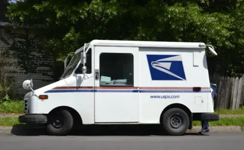 A white USPS mail delivery truck is parked on the side of a street. The vehicle features the USPS logo and web address on its side. There is foliage in the background and part of a person is barely visible near the back of the truck.