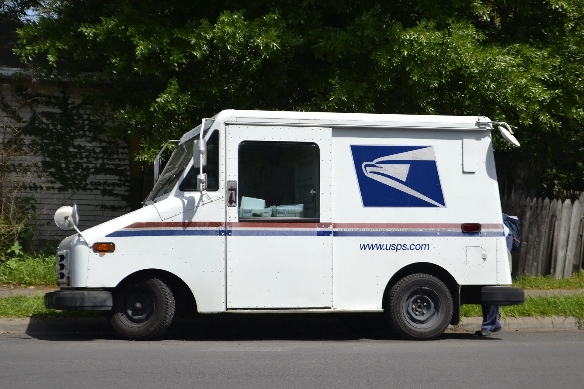 A white USPS mail delivery truck is parked on the side of a street. The vehicle features the USPS logo and web address on its side. There is foliage in the background and part of a person is barely visible near the back of the truck.