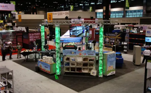 An overhead view of a convention booth featuring displays with colorful lights, promotional banners, and various products on shelves. A large screen is mounted at the top. Attendees are visible around the booth in a spacious exhibition hall.