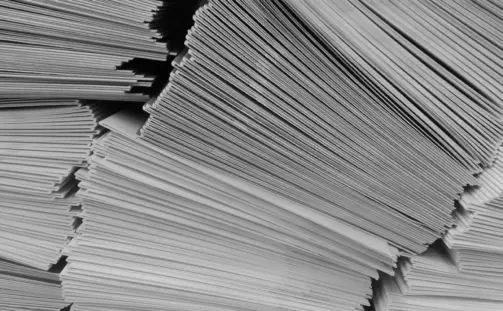 A black and white photo captures several tall stacks of paper sheets, neatly arranged. The papers create a pattern of lines and shadows, suggesting organized, voluminous documentation—perhaps alluding to the USPS August Rate Change preparation.