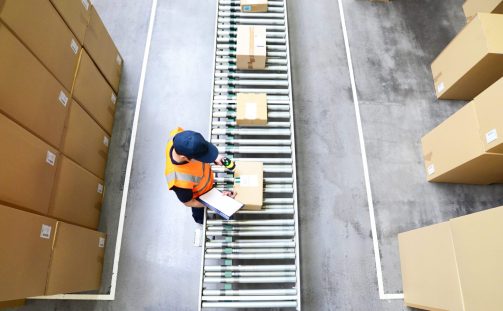 A warehouse worker in an orange vest and cap stands beside a conveyor belt with cardboard boxes, clipboard and scanner in hand. Surrounded by stacks of boxes on the floor, they are diligently preparing for Q4's demands.