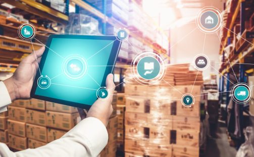 Person holding a tablet in a warehouse, featuring digital icons representing logistics and inventory management. Stacked pallets and shelves filled with boxes surround the scene, highlighting technology's role in modern supply chain operations.