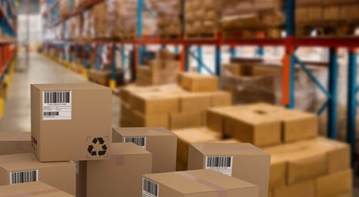 Stacks of cardboard boxes with barcodes are in the foreground of a large warehouse. Shelves filled with more boxes line the background, extending into the distance. The scene suggests an organized storage or distribution facility.