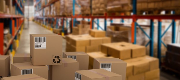 Stacks of cardboard boxes with barcodes are in the foreground of a large warehouse. Shelves filled with more boxes line the background, extending into the distance. The scene suggests an organized storage or distribution facility.