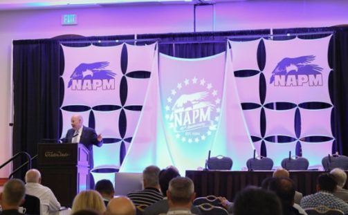 Post Master General Louis DeJoy addresses an audience at the NAPM conference, where the stage is adorned with the organization's logo and decorative panels. Attendees are seated, eagerly facing the stage in a well-appointed conference room.