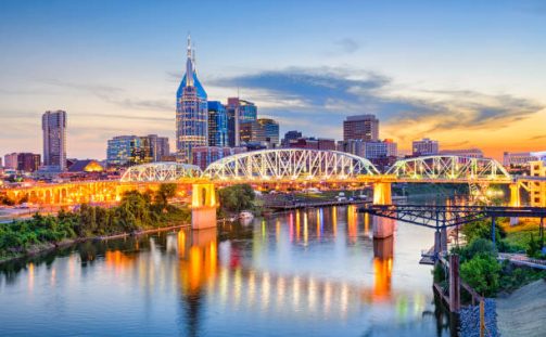 A vibrant city skyline at dusk, featuring illuminated skyscrapers and a prominent, arched bridge reflecting on a calm river. The sky transitions from orange to blue, adding a warm glow to the scene.