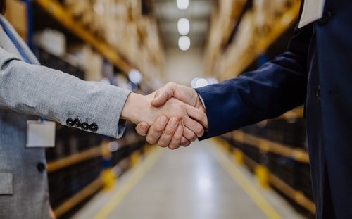 Two people shaking hands in a warehouse aisle, surrounded by shelves filled with boxes. Both are dressed in business attire, with one wearing a light gray suit and the other a dark blue suit. The background is slightly blurred.