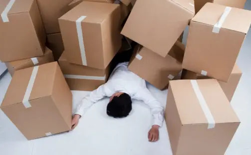 A person in a white shirt lies on the floor, partially covered by a pile of large cardboard boxes. The scene suggests an accidental collapse of the boxes around them. The floor and walls are white, highlighting the boxes and the person.