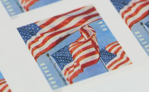 Close-up of U.S. postage stamps featuring the American flag waving in the wind against a blue sky. The word 