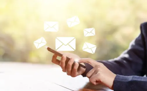 A person is using a smartphone at a wooden table, with digital icons of envelopes floating above, symbolizing sending or receiving emails. The background is softly blurred with natural lighting.