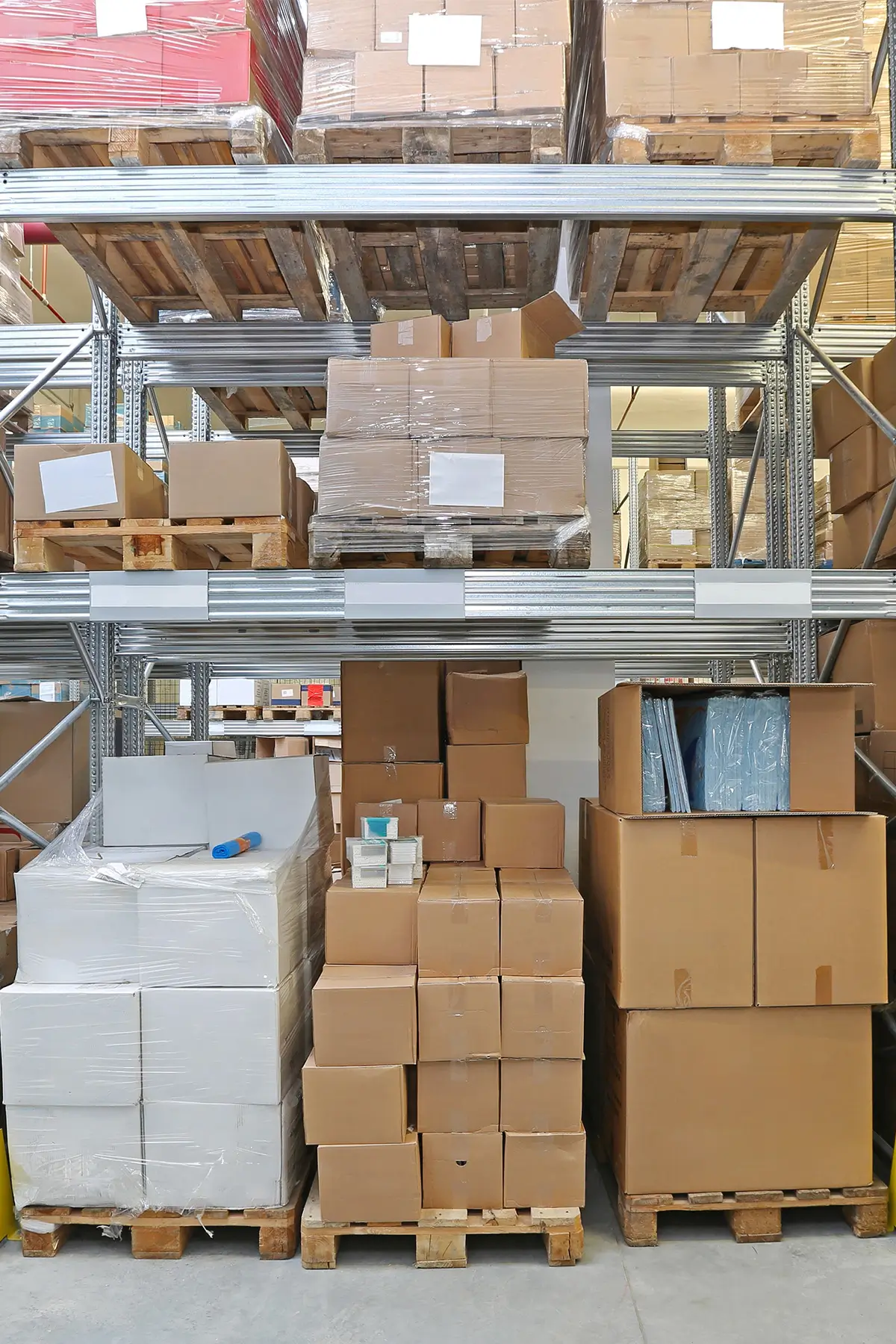 Stacks of cardboard boxes and pallets are organized on metal shelving in a warehouse. The boxes vary in size and are neatly arranged, with some wrapped in plastic. The floor is concrete, and the setting appears orderly and stocked.