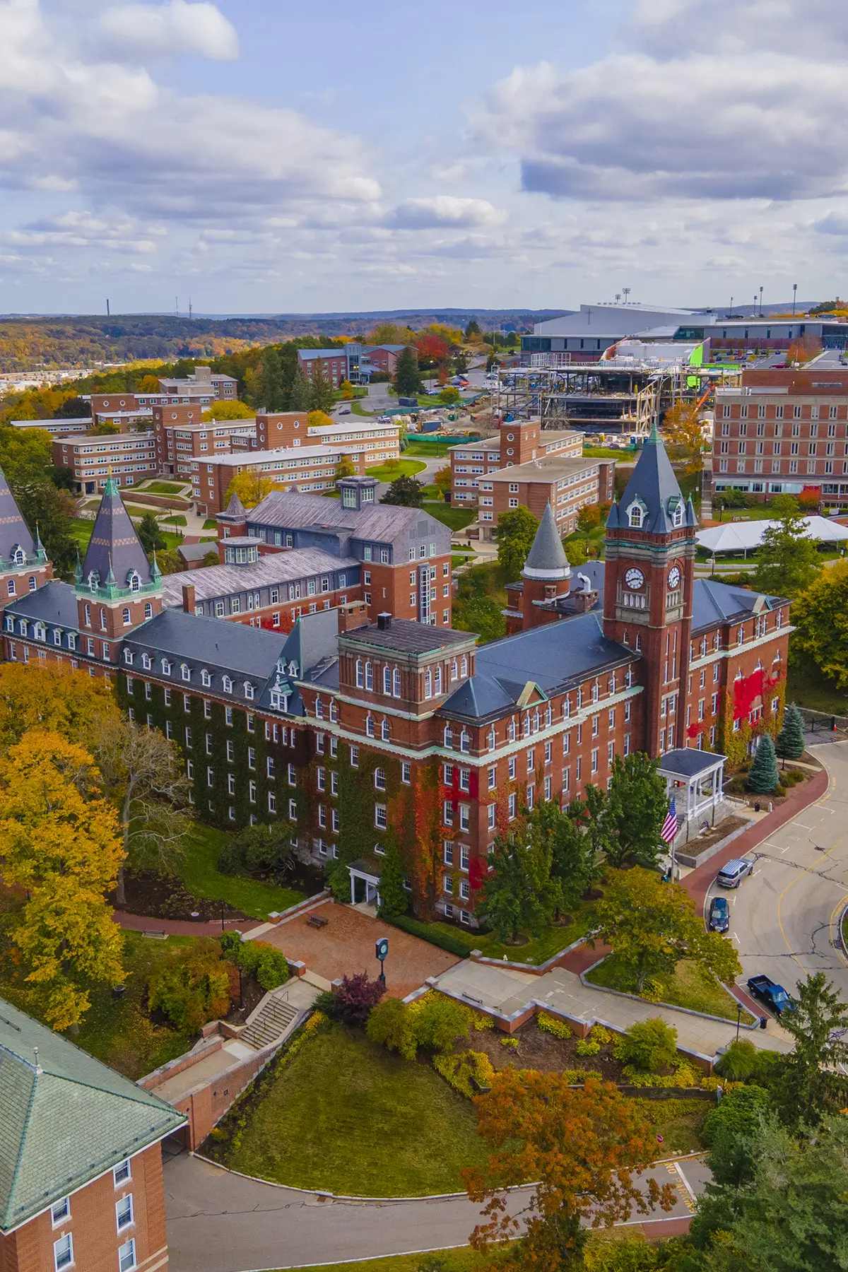 Aerial view of a large, historic campus with red-brick buildings featuring towers, set amidst autumn foliage. Paved pathways and roads wind through the property, with distant bleachers and a stadium visible against a cloudy sky.