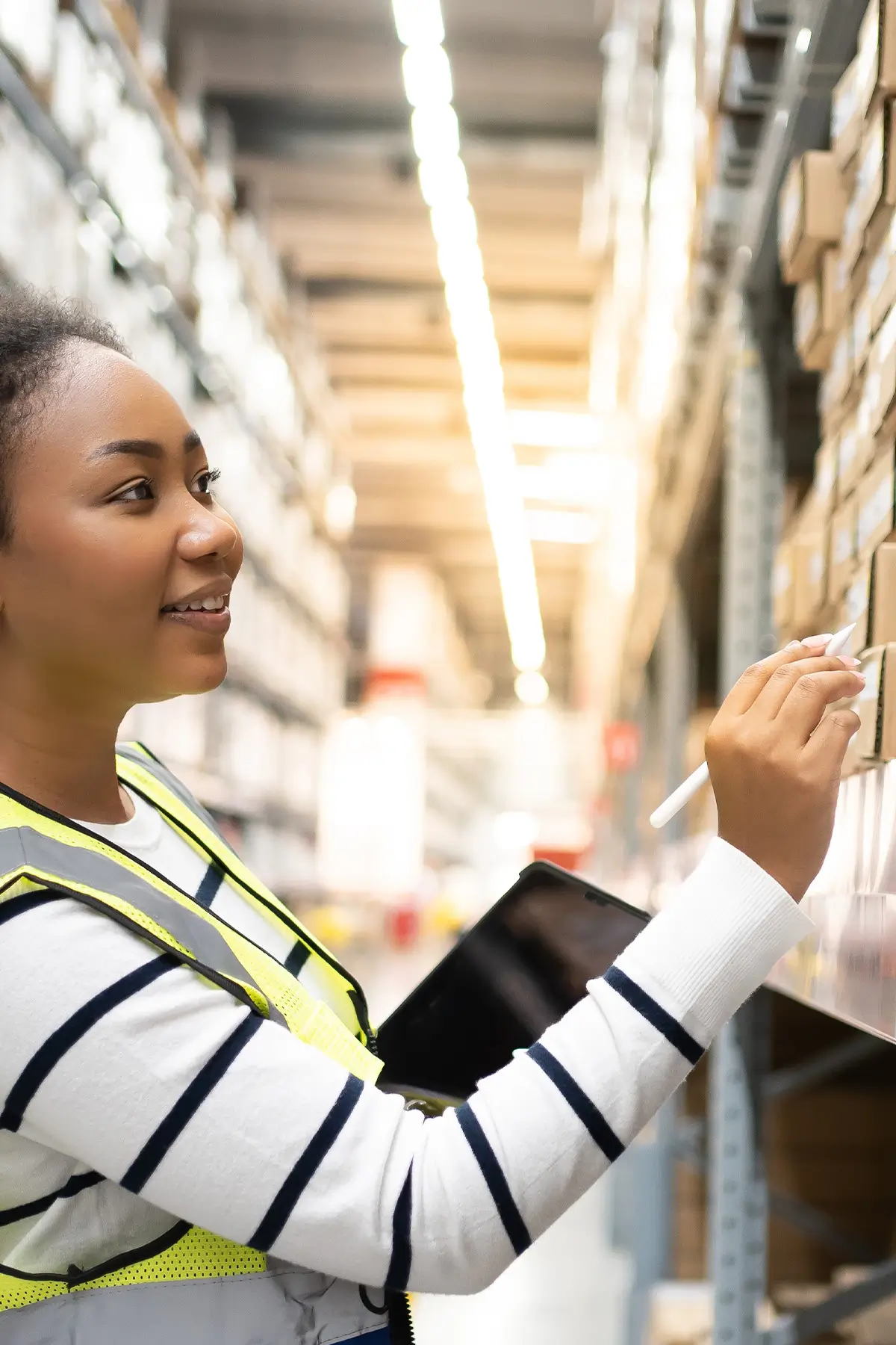 A woman wearing a striped shirt and a high-visibility vest is holding a tablet and writing on a box in a warehouse aisle filled with shelves of boxes. She appears focused and is smiling slightly.