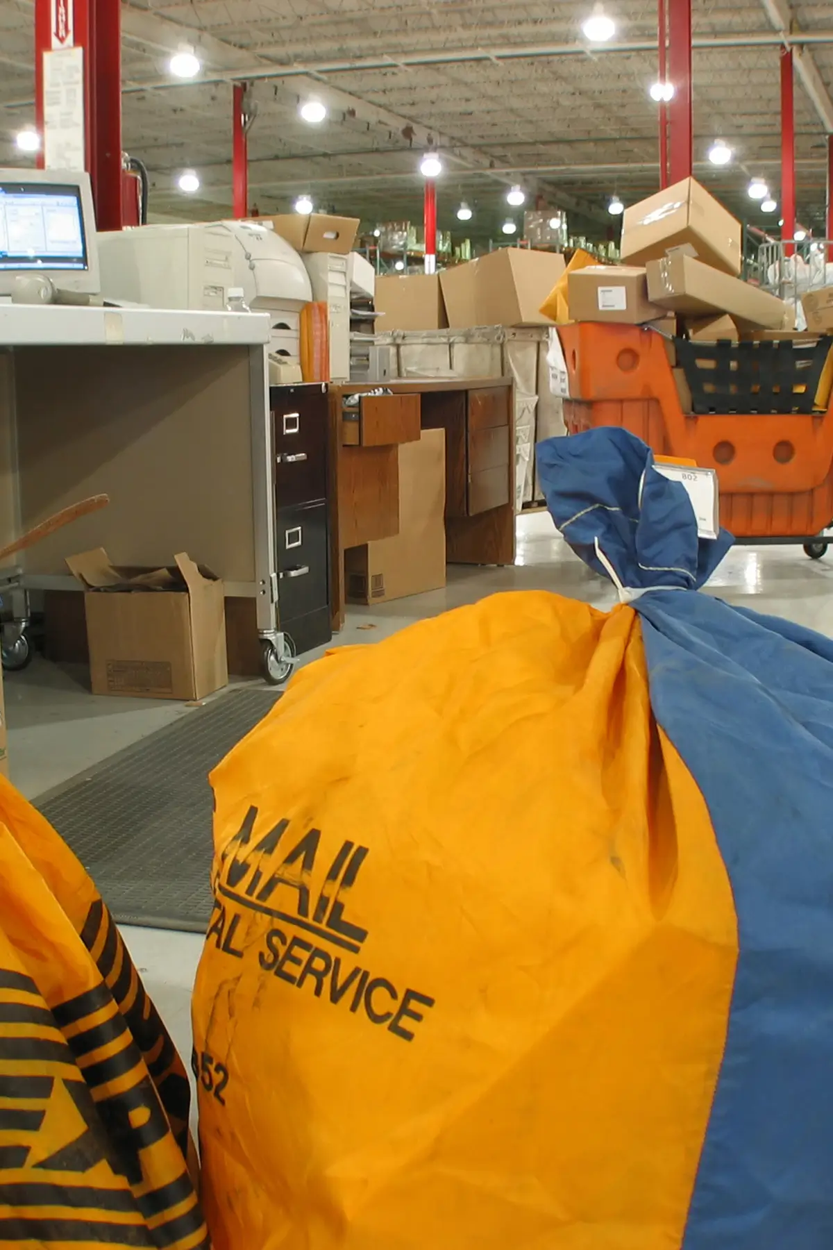 A mail processing facility with large yellow and blue postal bags in the foreground. In the background, there are bins, boxes, and envelopes, along with a computer workstation under bright overhead lights.