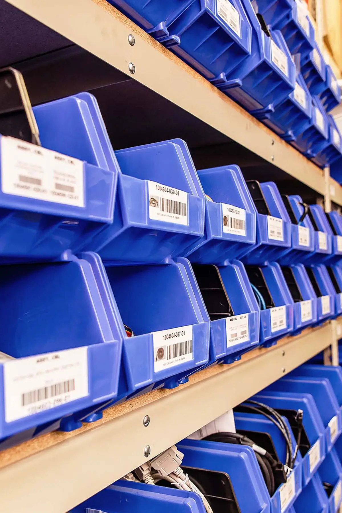 Blue plastic bins with labels are neatly arranged on shelves in a storage area. Some bins contain various small items, and barcodes are visible on the labels. The shelves are made of light-colored wood.