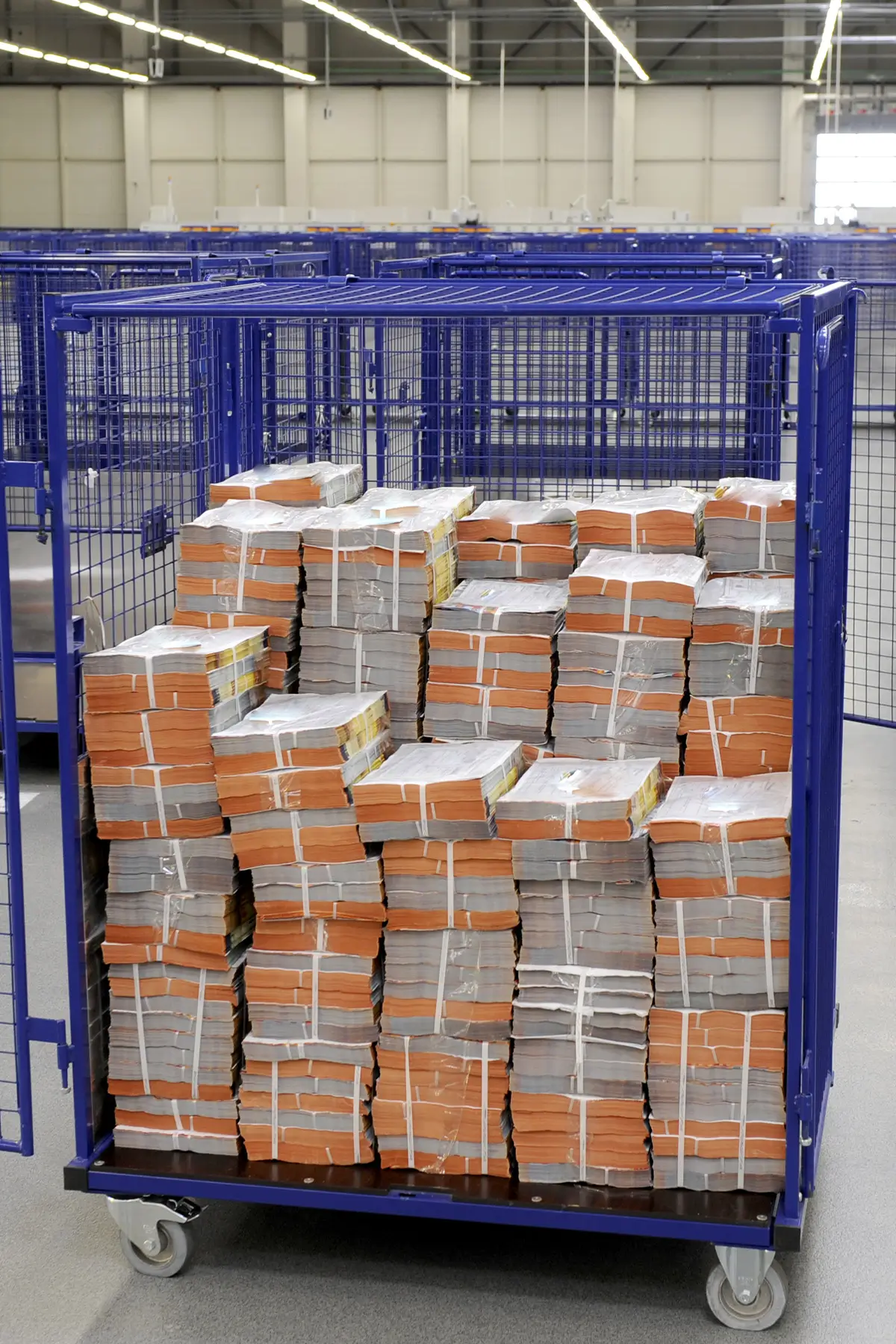 A large blue metal cart on wheels is filled with stacks of bundled paper, wrapped with white straps. The cart is situated in a spacious room with additional similar carts visible in the background.