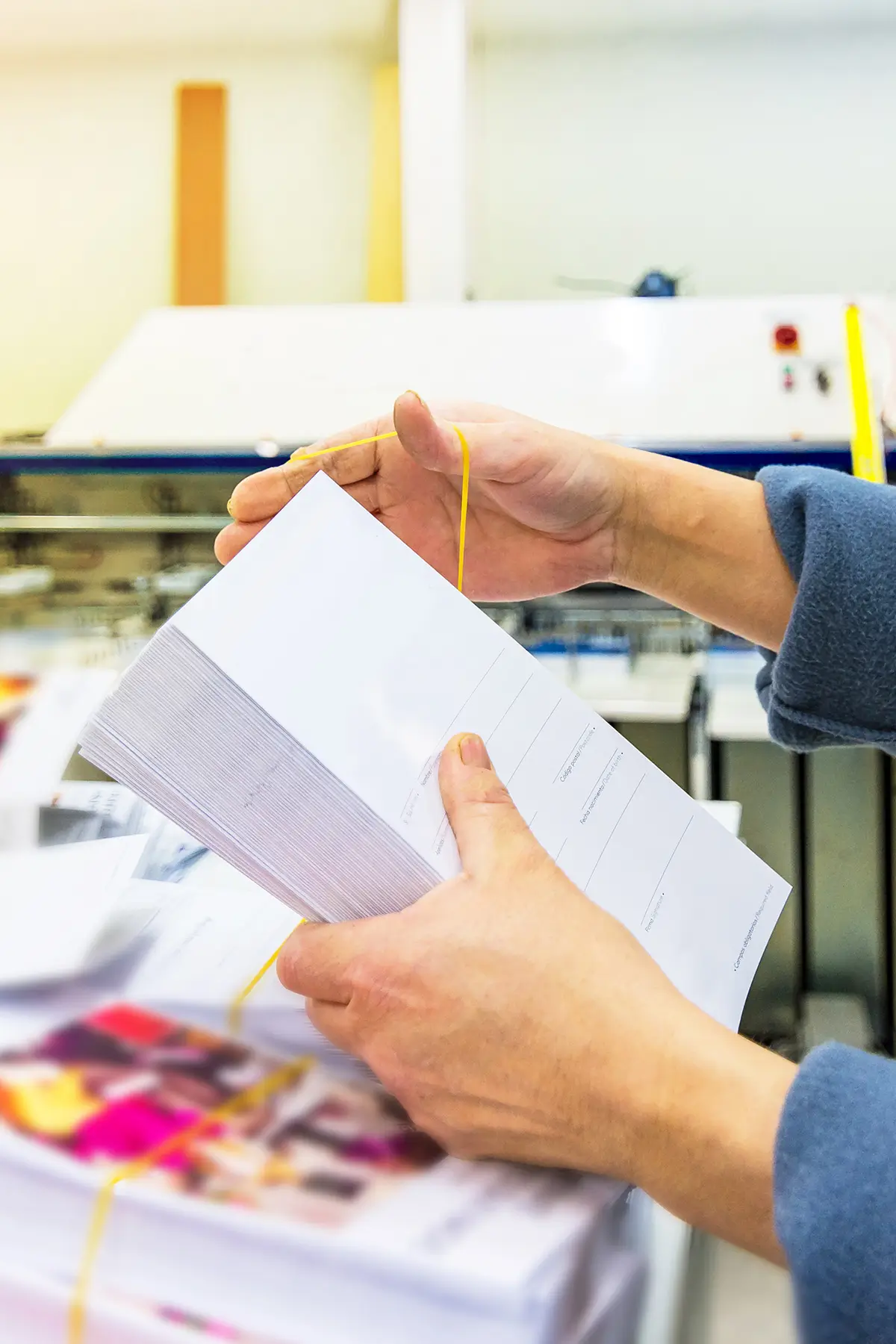 Hands holding a stack of papers secured with a yellow rubber band, in an office or print shop setting. Various colorful printed materials are visible in the background.