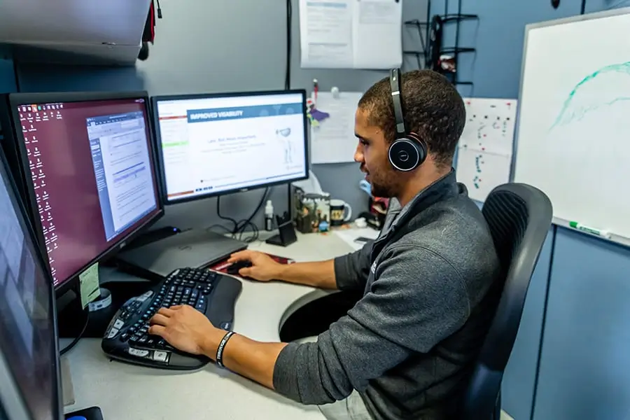 A person wearing headphones is sitting at a desk with three computer monitors. They are focused on the screens, which display documents and charts in a workspace with grey walls and a whiteboard nearby.