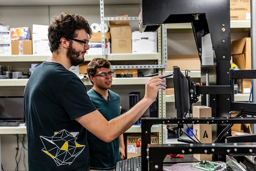 Two men are working on a computer monitor in a workshop filled with shelves of tools and boxes. One man is adjusting the settings on a screen while the other observes. Both appear focused and engaged in the activity.