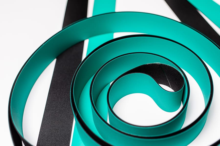 A close-up of coiled, flat bands in black and turquoise. The bands create a spiral effect on a white background, with additional flat strips in the same colors lying nearby.