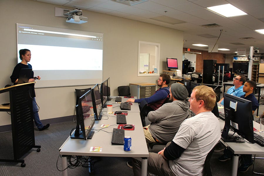 A person is giving a presentation to a small group seated at computer desks in a classroom. The presenter stands near a screen displaying a website. Attendees are focused on the presentation.