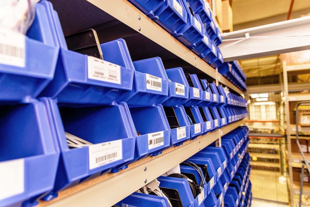 Rows of blue plastic bins on shelves in a warehouse, each labeled with barcodes. The bins contain various small components, indicating organized storage. The bright lighting highlights the orderly arrangement of the shelves.