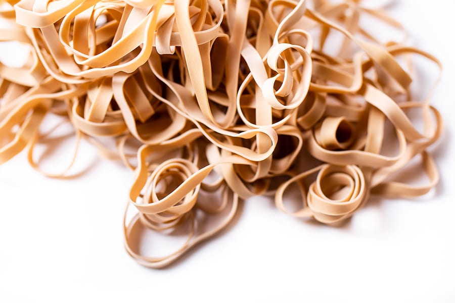 A pile of beige rubber bands tangled together, set against a plain white background. The bands overlap in various directions, creating a complex and textured pattern.