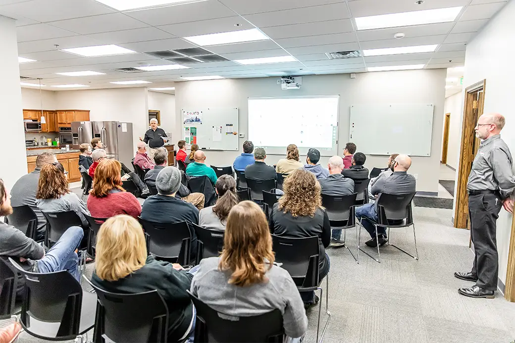 A group of people in a classroom setting sit on chairs facing a projector screen, where a person at the front is presenting. Another person stands near the side of the room. The setting appears informal, with a kitchen area visible in the background.