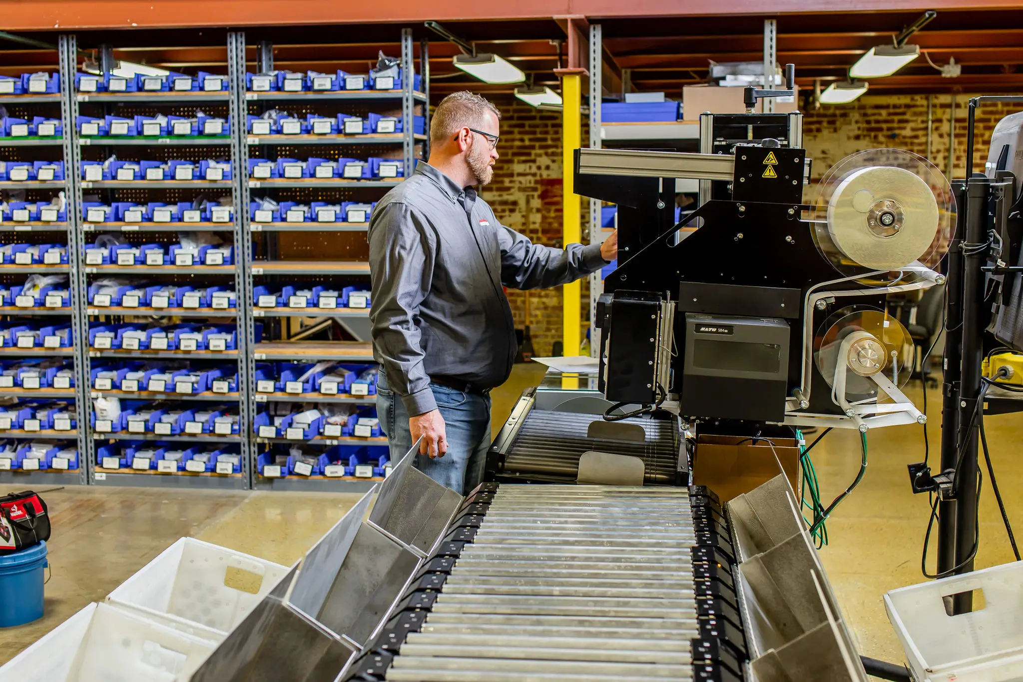 A person operates machinery in a warehouse setting. The area is organized with shelves filled with small boxes. The individual is focused on the controls, wearing a gray shirt and jeans, standing by a conveyor belt.