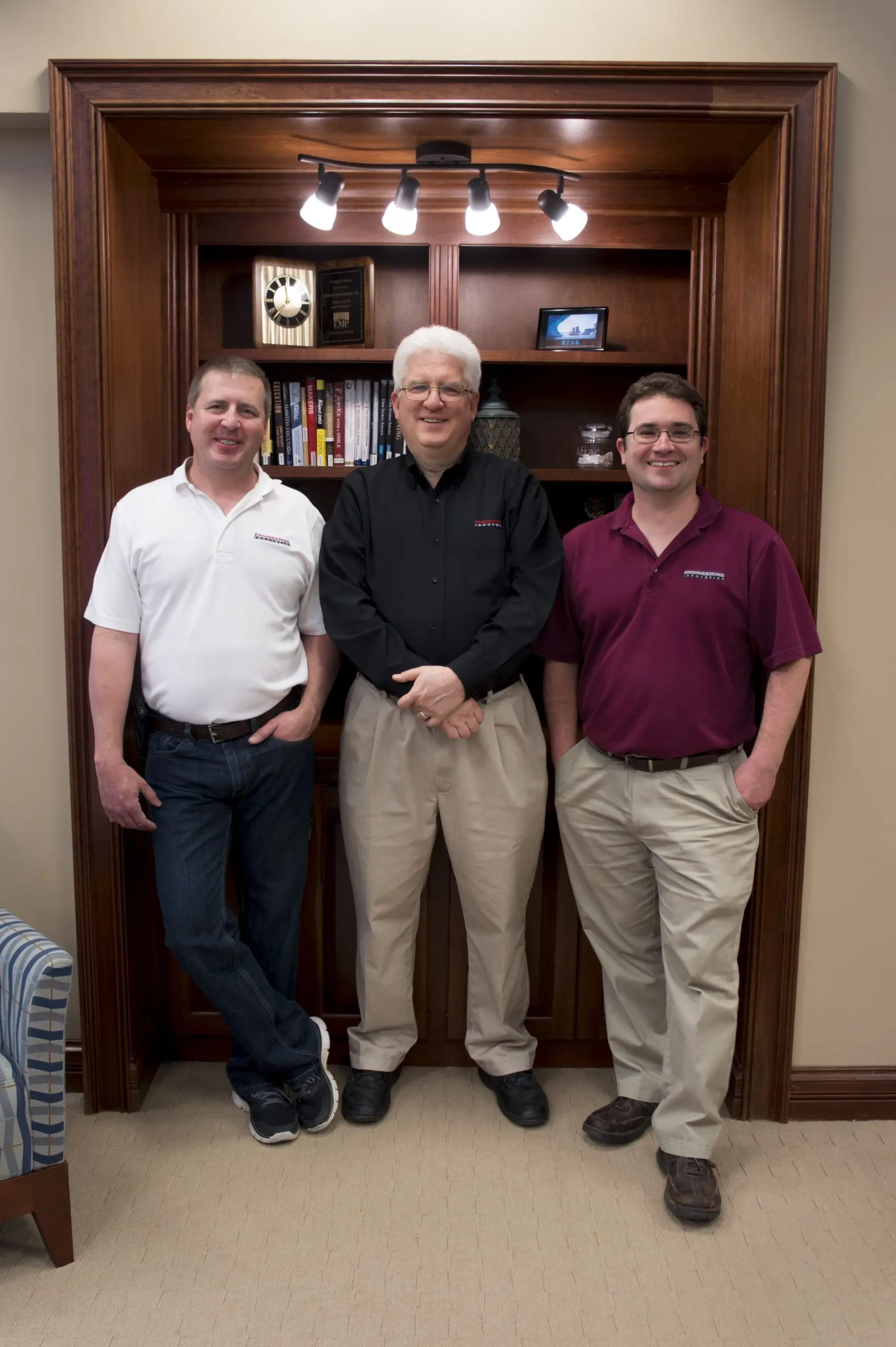 Three men stand in front of a wooden bookshelf with books and decorations. They wear casual shirts, each in different colors: white, black, and maroon. The setting appears to be a cozy office or study.