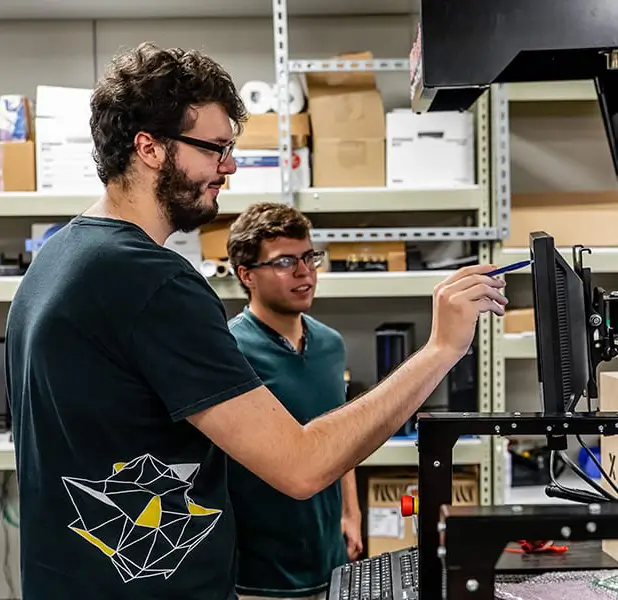 Two people with beards are working in a workshop. One is pointing at a computer screen on a machine, while the other stands behind him. Shelving with boxes is visible in the background. They wear glasses and casual dark t-shirts.