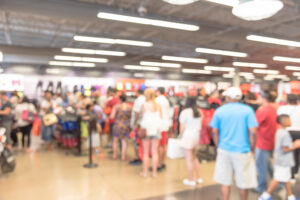 A blurred image of a busy clothing store with many people browsing and shopping. The interior is bright with overhead lights and various clothes displays.