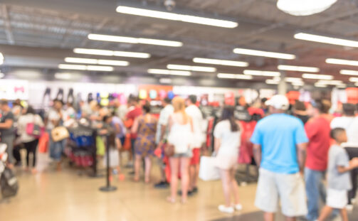 A blurred image of a busy clothing store with many people browsing and shopping. The interior is bright with overhead lights and various clothes displays.