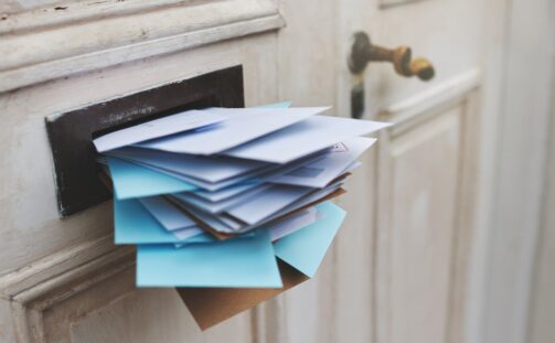 A door with a full letterbox overflowing with a stack of white and blue envelopes, suggesting a large amount of mail has accumulated. The door is cream-colored with a bronze handle.