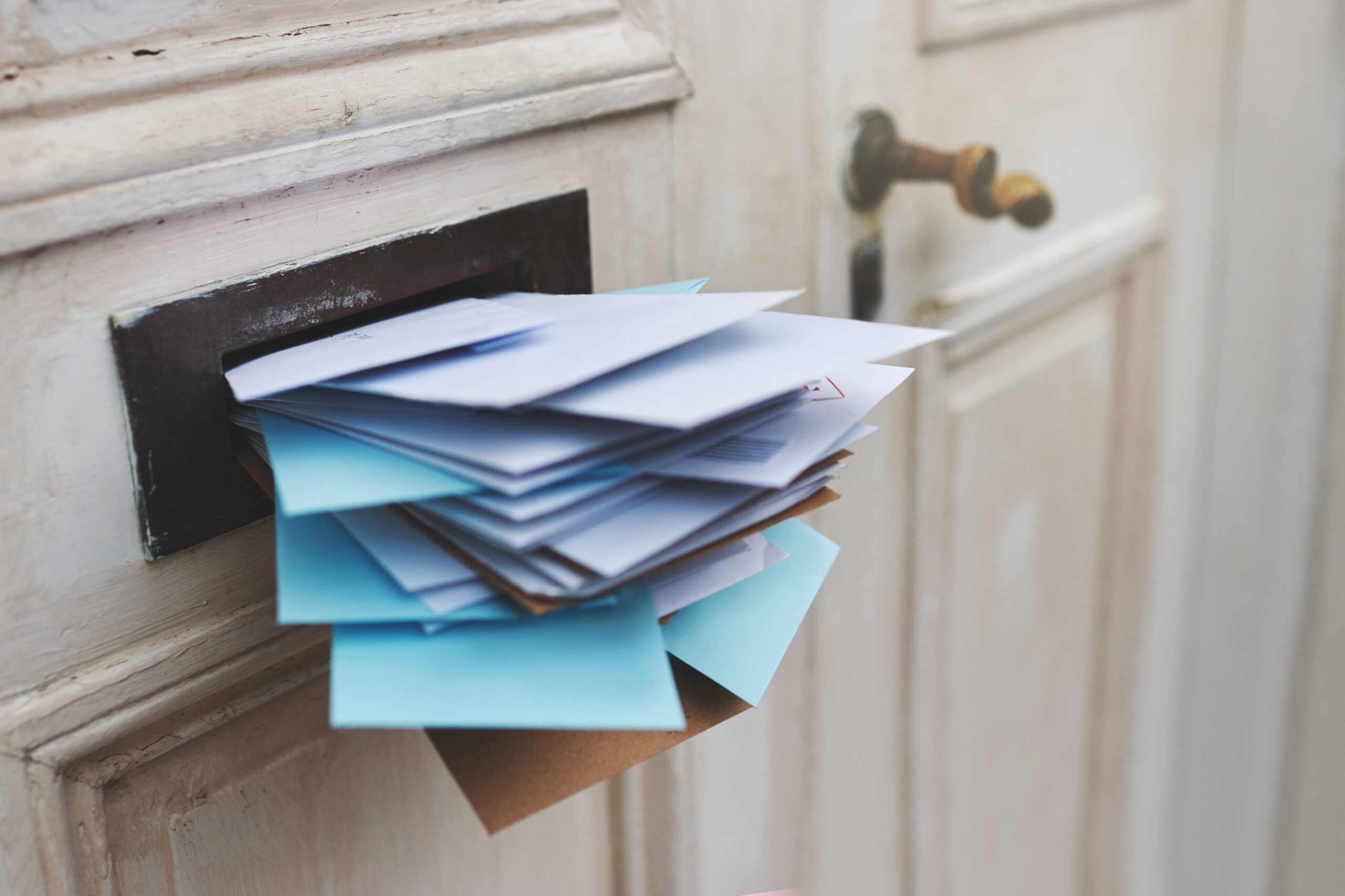 A door with a full letterbox overflowing with a stack of white and blue envelopes, suggesting a large amount of mail has accumulated. The door is cream-colored with a bronze handle.