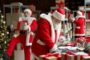 Three people dressed as Santa are in a bustling warehouse, navigating the holiday mail surge. One checks a list at a desk with a laptop, another carries a gift, and the third is placing it on a shelf. A beautifully decorated Christmas tree adds to the festive chaos of wrapped gifts and shelves.