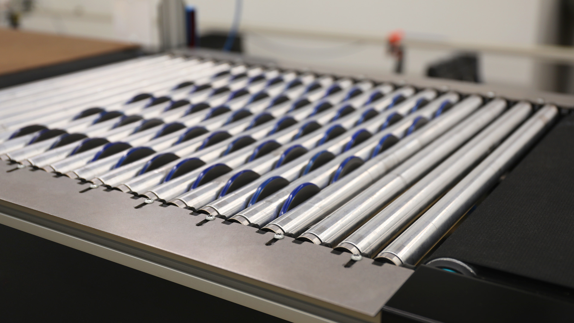 Conveyor rollers in a manufacturing setting. The metallic rollers are aligned horizontally and mounted on a frame, suggesting a system for transporting items. The background features blurred machinery components.