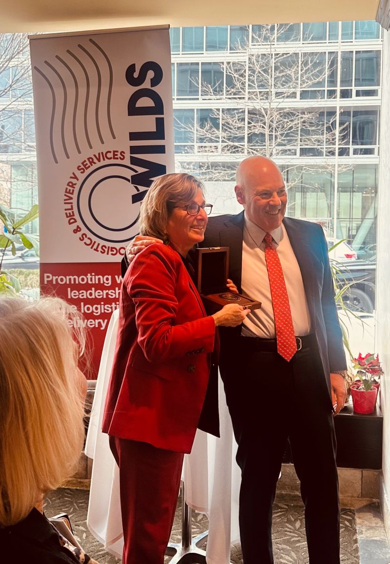 A woman in a red suit holds an award next to a man in a suit with a red tie. They are standing in front of a banner that reads "WILDS Women in Logistics & Delivery Services." The background shows a window with a view of buildings.