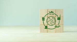 Wooden blocks depict a green circular diagram with a globe at the center. Surrounding the globe are icons for a trash bin, a shopper, a truck, and a factory, symbolizing recycling and environmental sustainability—key elements in logistics trends 2025.