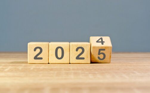 Wooden blocks on a table display the year 2024, with the last block being turned to reveal 2025, symbolizing upcoming logistics trends. The background is plain and gray.