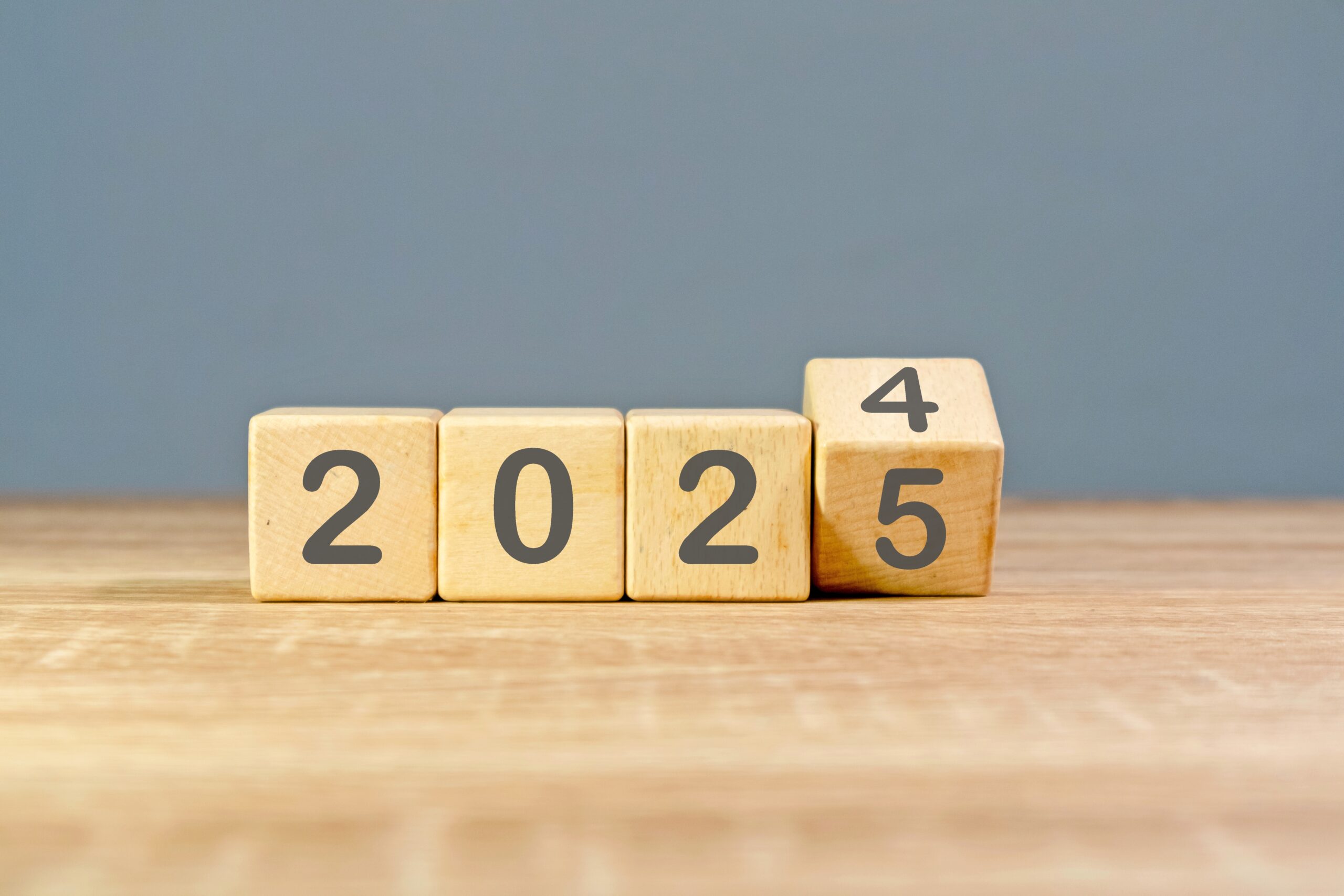 Wooden blocks on a table display the year 2024, with the last block being turned to reveal 2025, symbolizing upcoming logistics trends. The background is plain and gray.