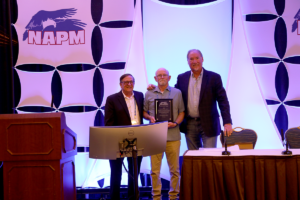 Three men stand on stage at a podium, one proudly holding a plaque. The backdrop features the iconic NAPM 2025 logo with an eagle. In the foreground, a monitor and tables complete the setting.